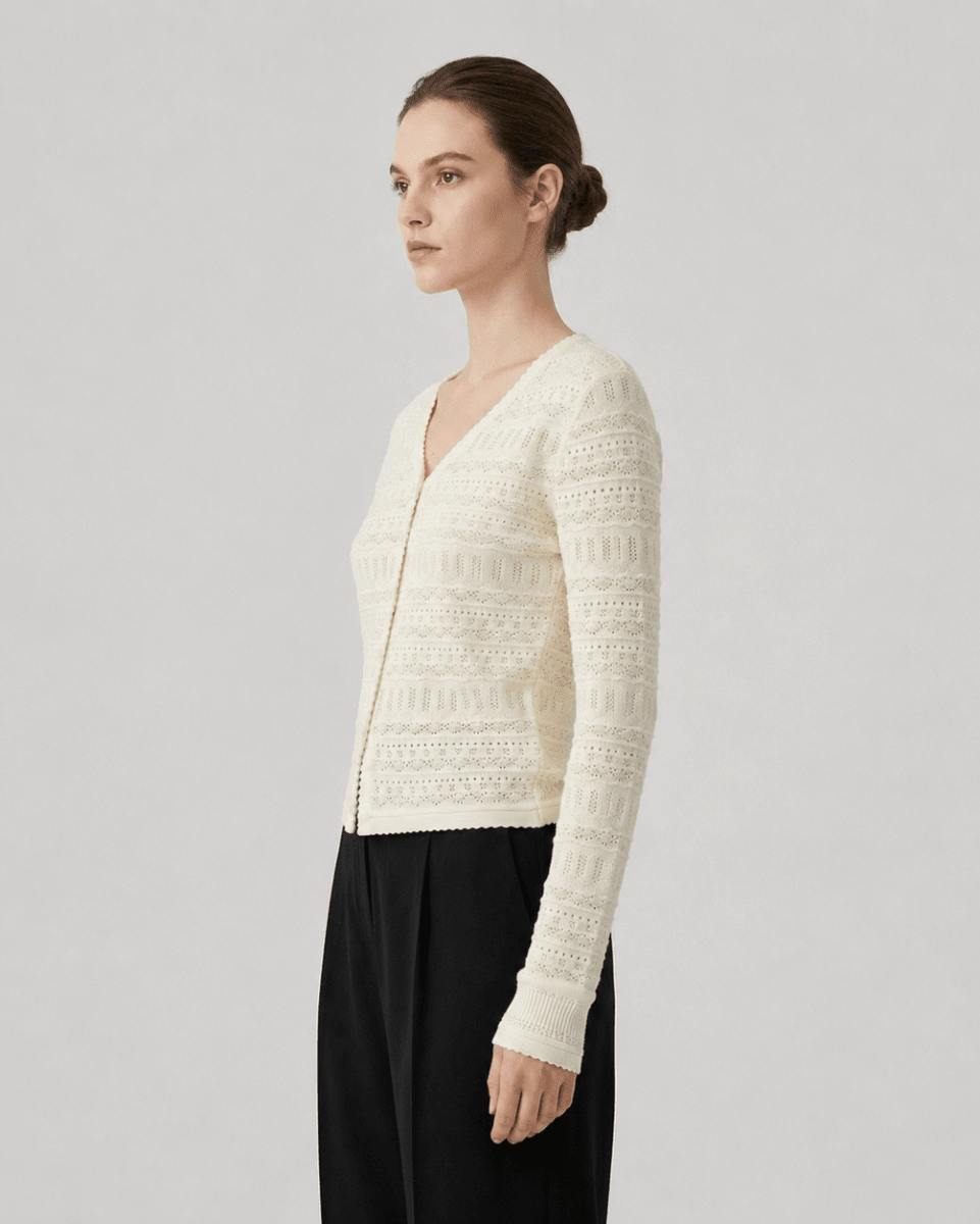 A woman modeling a cream-colored, long-sleeve crochet knit cardigan with a V-neck and front zipper, paired with black wide-leg trousers, against a solid light gray studio background.