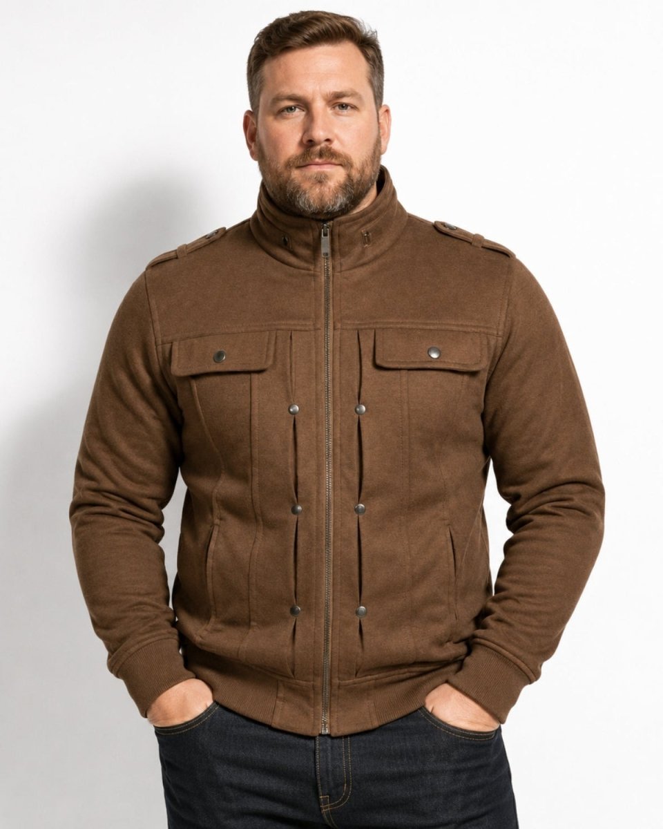 A man wearing a brown zip-up jacket with a high collar, buttoned chest pockets, and snap-front detailing, paired with dark jeans, standing against a plain white studio background.