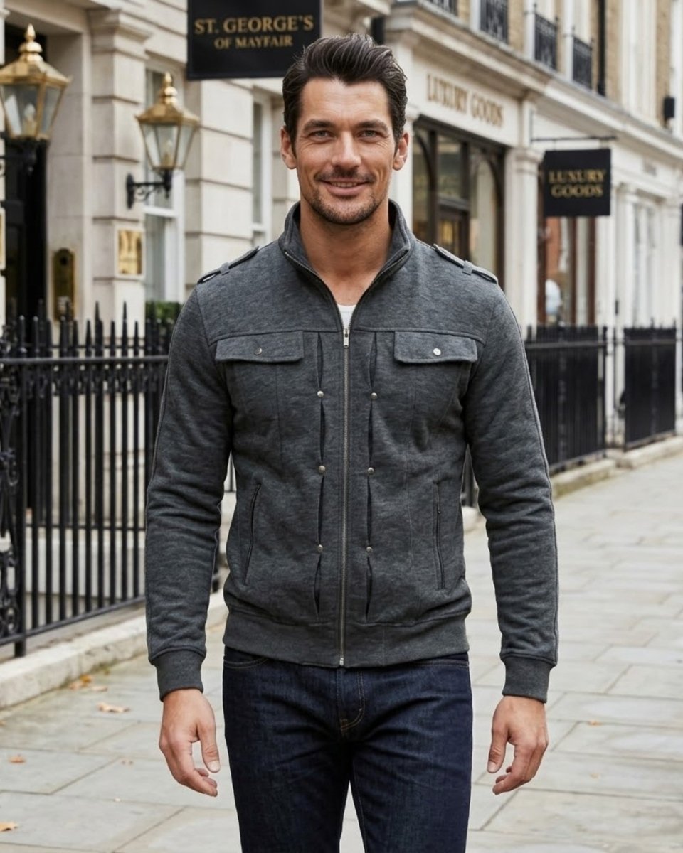 A man wearing a dark gray zip-up jacket with flap chest pockets and dark blue jeans, standing on a city sidewalk in front of a building with storefronts.