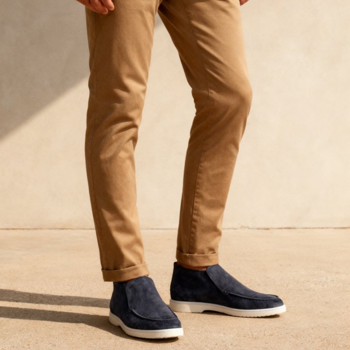 Kouvr suede ankle loafer boots with tan chino pants.
Side view of black Italian suede slip-on boots with a contrasting white sole, worn by a model.