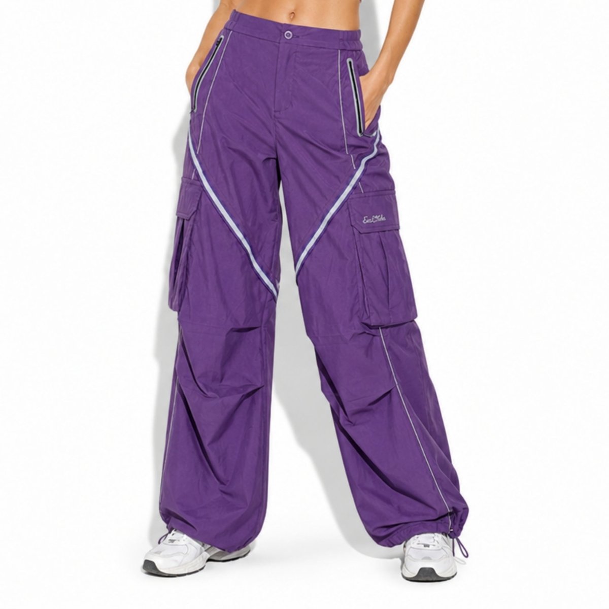 Front view of model wearing Kouvr oversized purple parachute cargo pants with white contrast piping, styled with a white ribbed crop top and chunky sneakers.