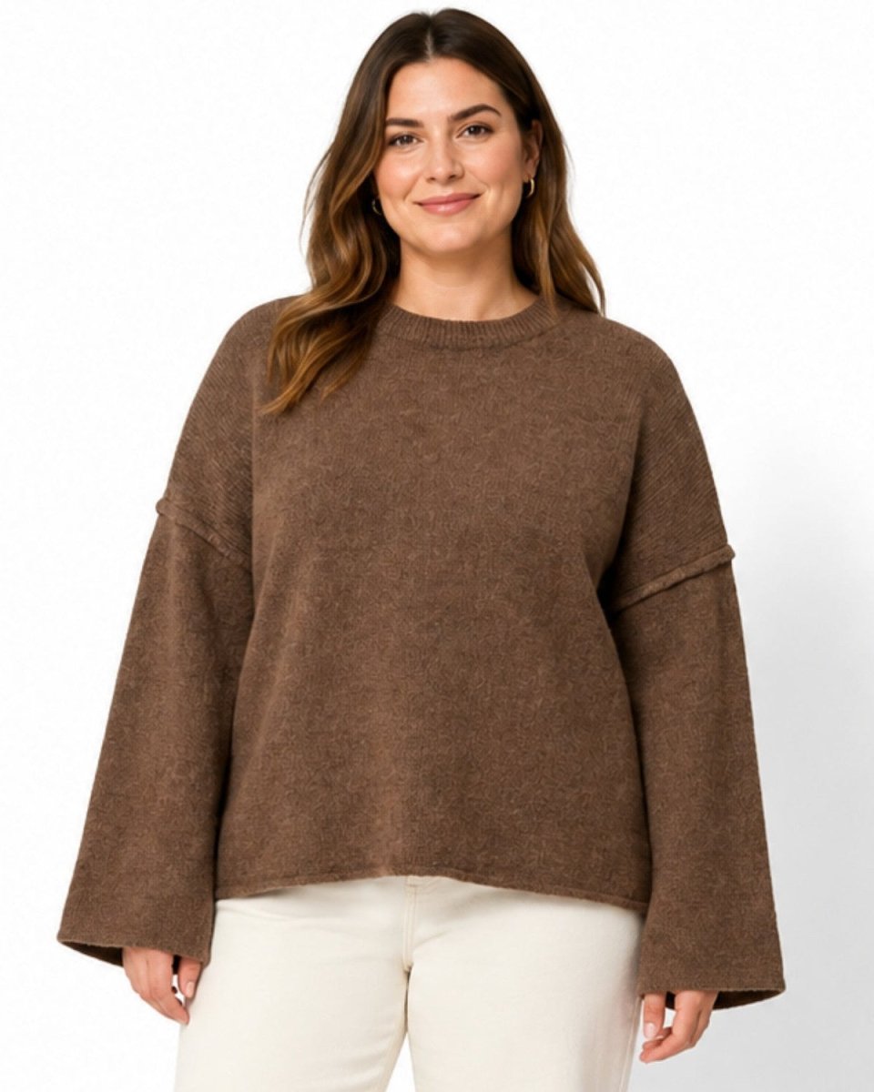 A woman modeling a brown knit sweater with wide bell sleeves and a crew neck, paired with off-white pants, against a plain white studio background.