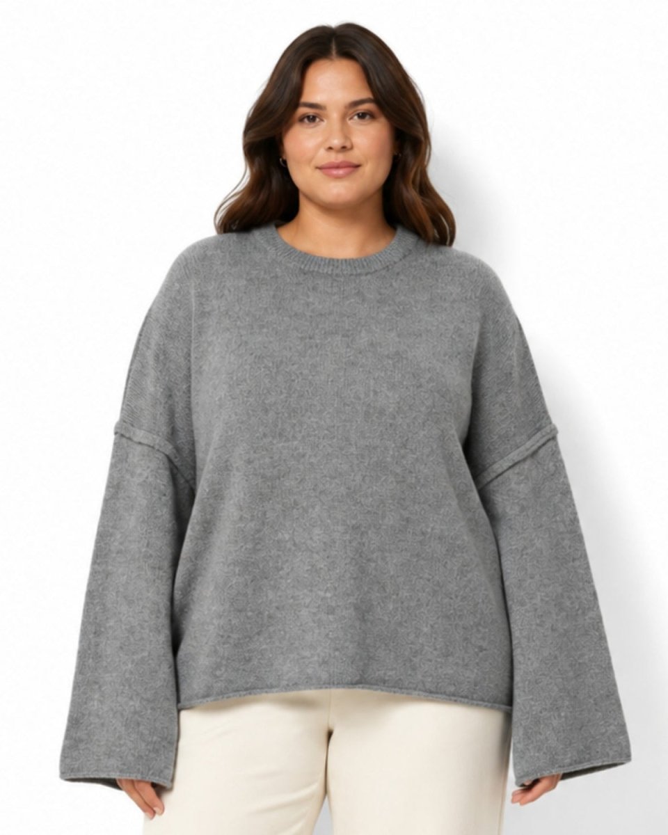 A woman modeling a heather gray oversized crewneck sweater with wide, slightly dropped long sleeves, paired with cream pants, against a plain white background.