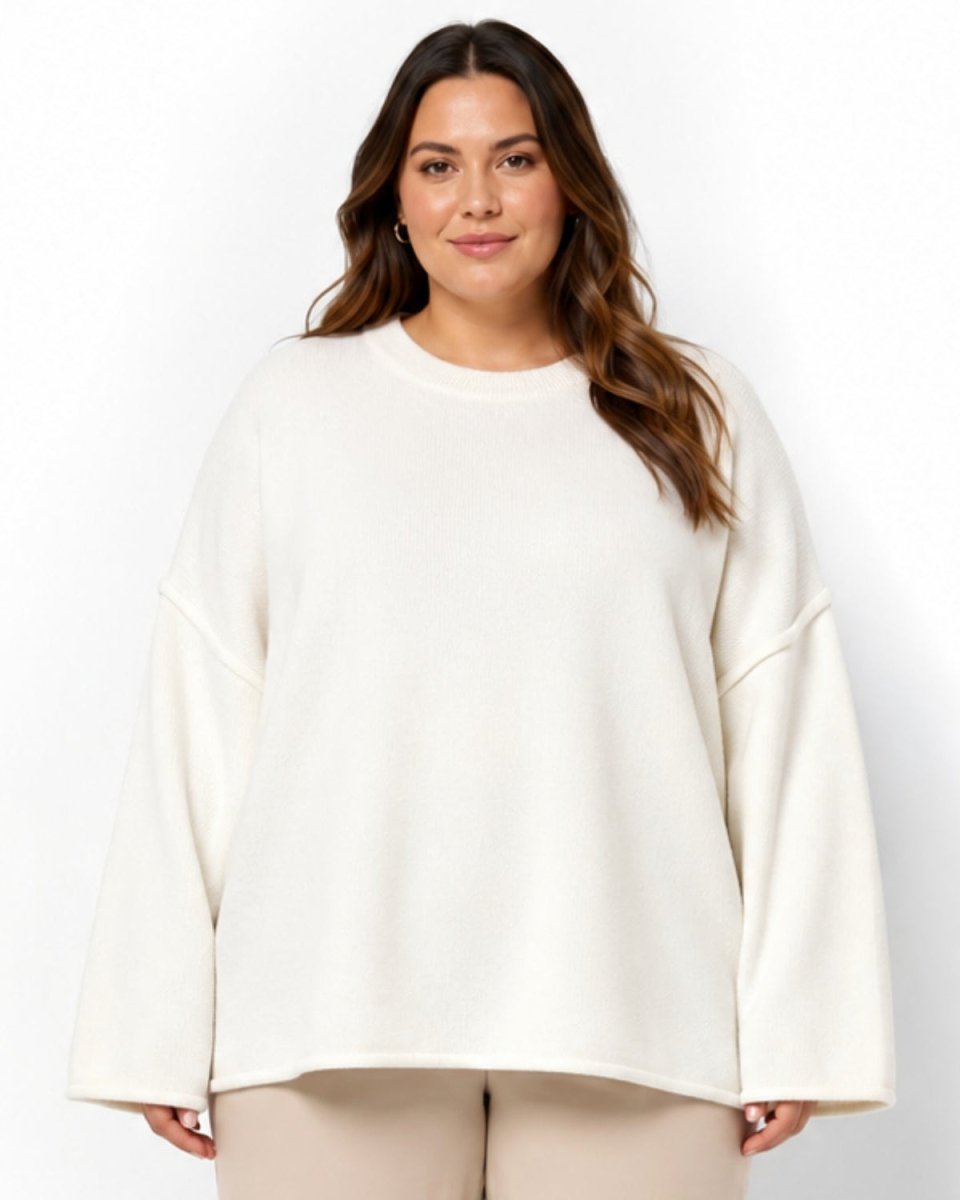 A woman modeling an oversized cream-colored crewneck sweatshirt with wide, slightly dropped-shoulder sleeves, paired with light beige pants, against a plain white studio background.