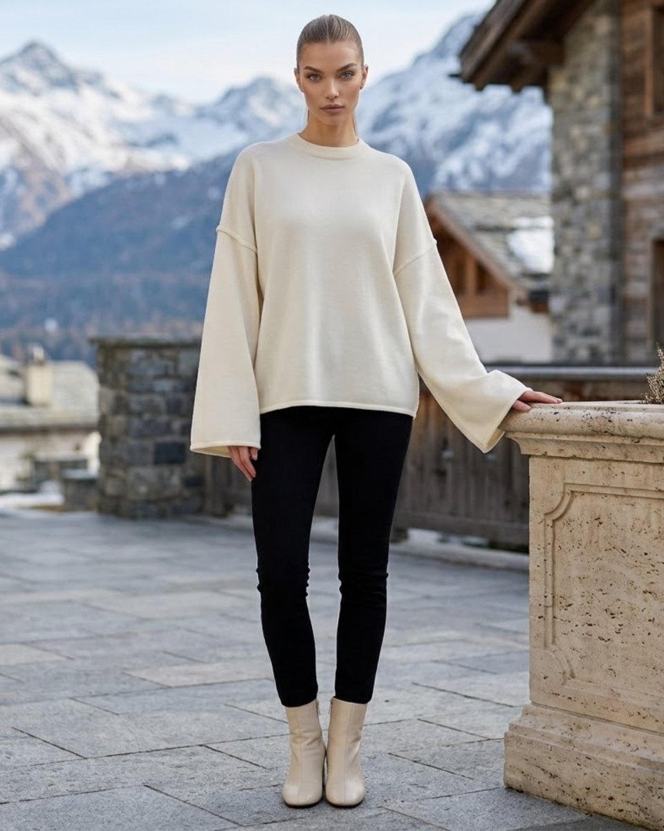 A woman stands outdoors in front of snowy mountains and stone buildings, wearing a cream knit sweater with dramatic bell sleeves, black skinny pants, and light beige ankle boots.