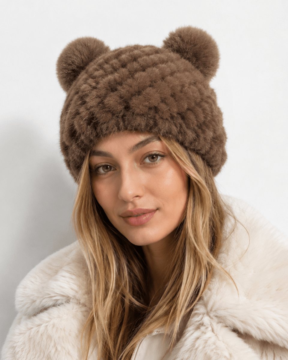 A woman wearing a brown fuzzy knit beanie with two pom-poms on top, paired with a cream faux-fur coat, against a plain light gray background.