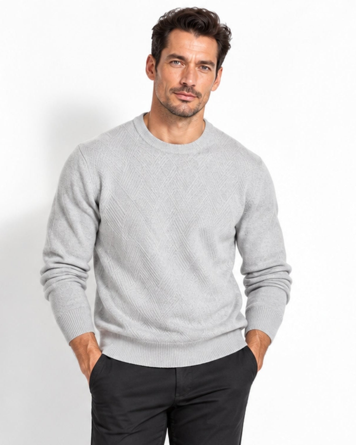 A man wearing a light gray textured knit crewneck sweater and dark trousers, standing against a plain white studio background.
