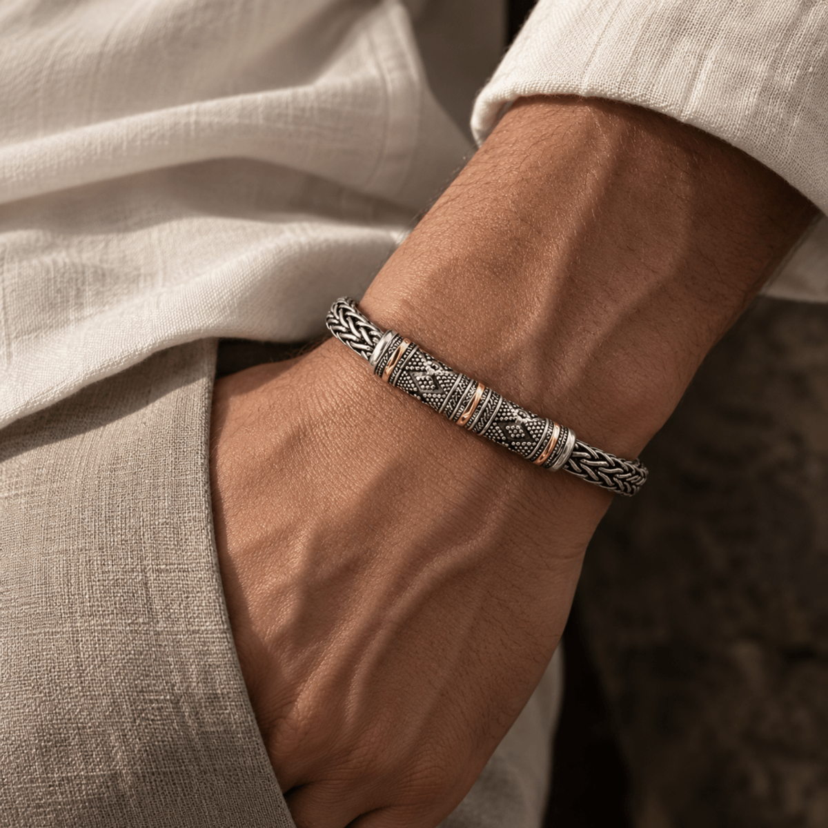 A close-up of a man's wrist wearing a textured silver-toned metal bracelet with geometric patterns and small gold accents, paired with a light beige linen shirt.