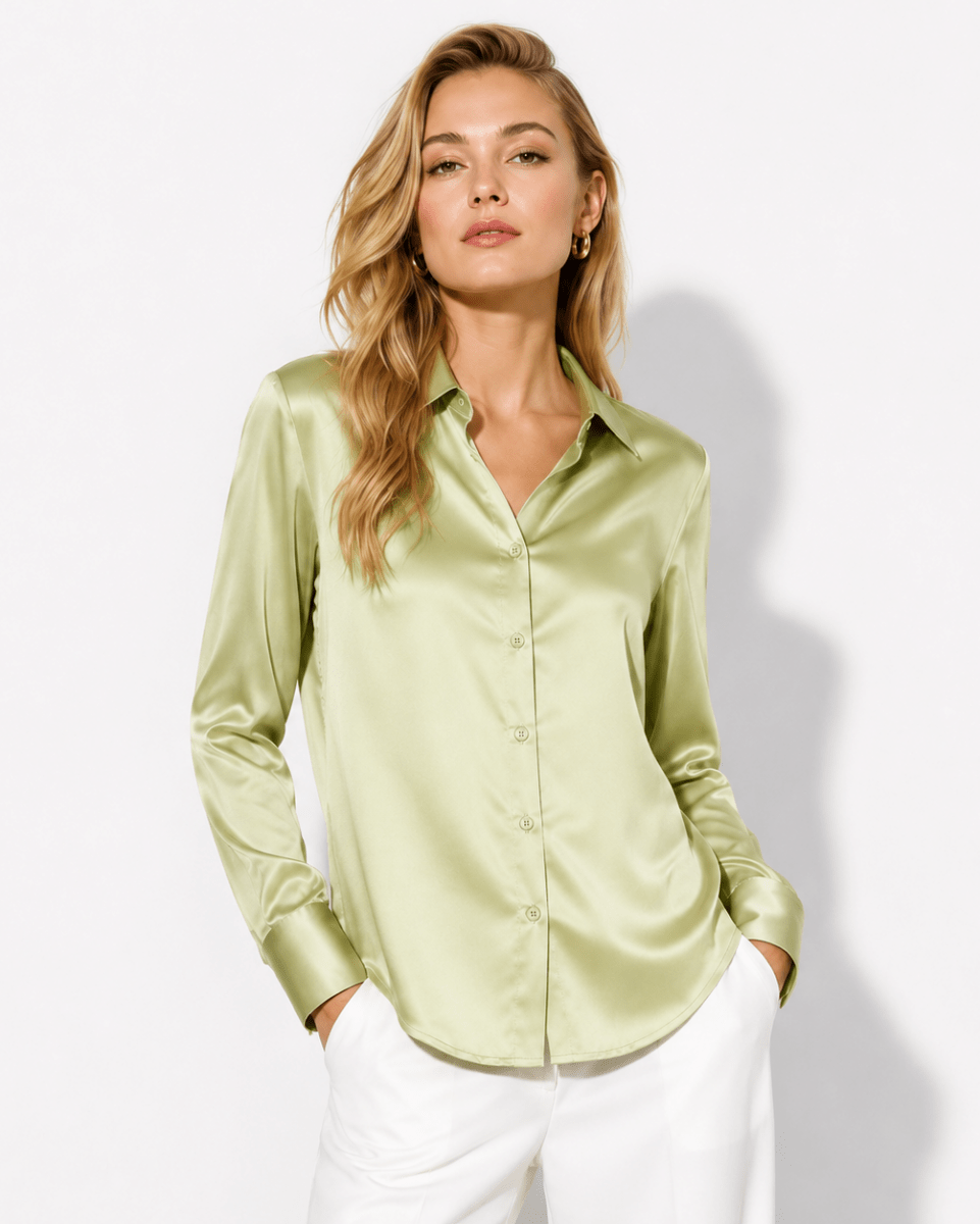 A woman modeling a light olive green satin button-down shirt with white pants, standing against a plain white studio background.