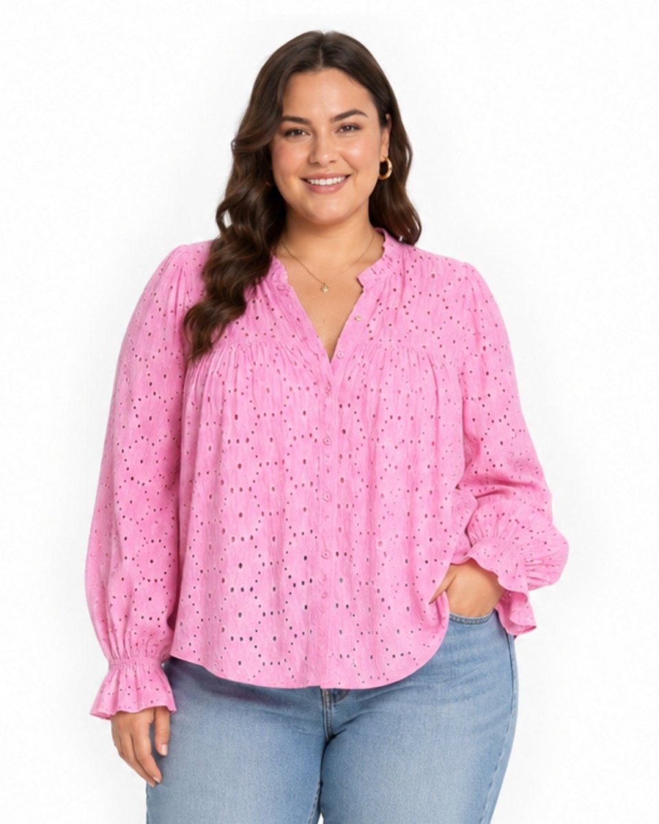 A woman wearing a pink eyelet long-sleeve blouse with a V-neck and ruffled cuffs, paired with light blue jeans, against a plain white background.
