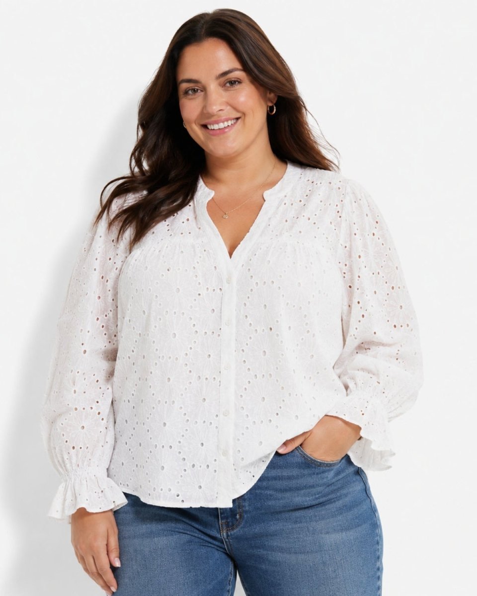 A woman wearing a white eyelet long-sleeve blouse with ruffled cuffs and a split neckline, paired with blue jeans, against a plain white studio background.