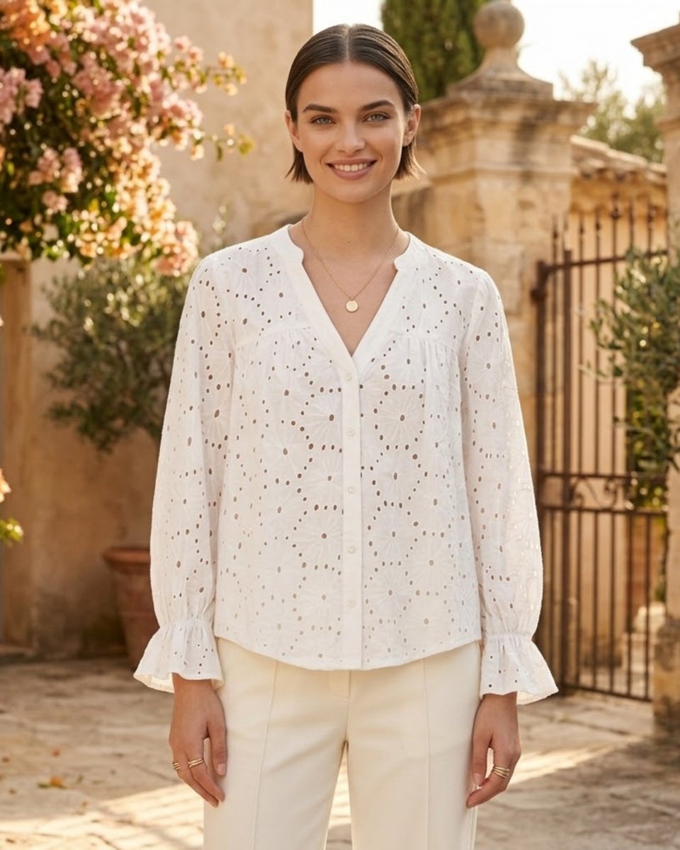 A woman wearing a white eyelet long-sleeve blouse with ruffled cuffs and a V-neckline, paired with cream trousers, standing in a sunlit outdoor courtyard with stone walls and pink flowers.