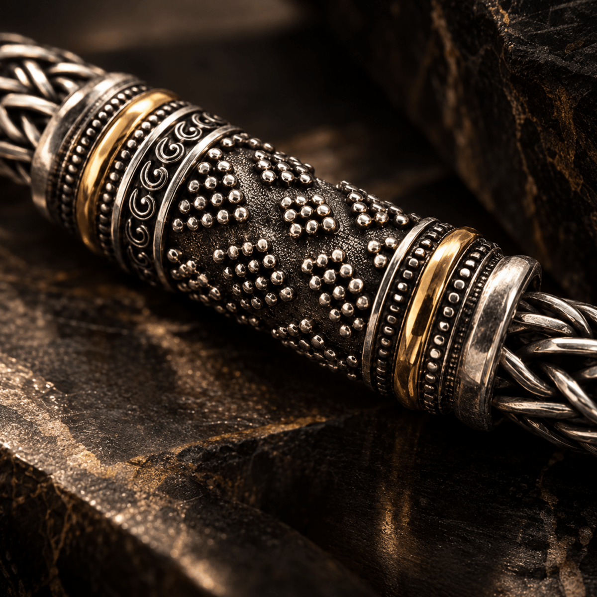 A close-up of an ornate braided metal cord or cable with decorative silver-toned and gold-toned bands, resting on dark textured stone.