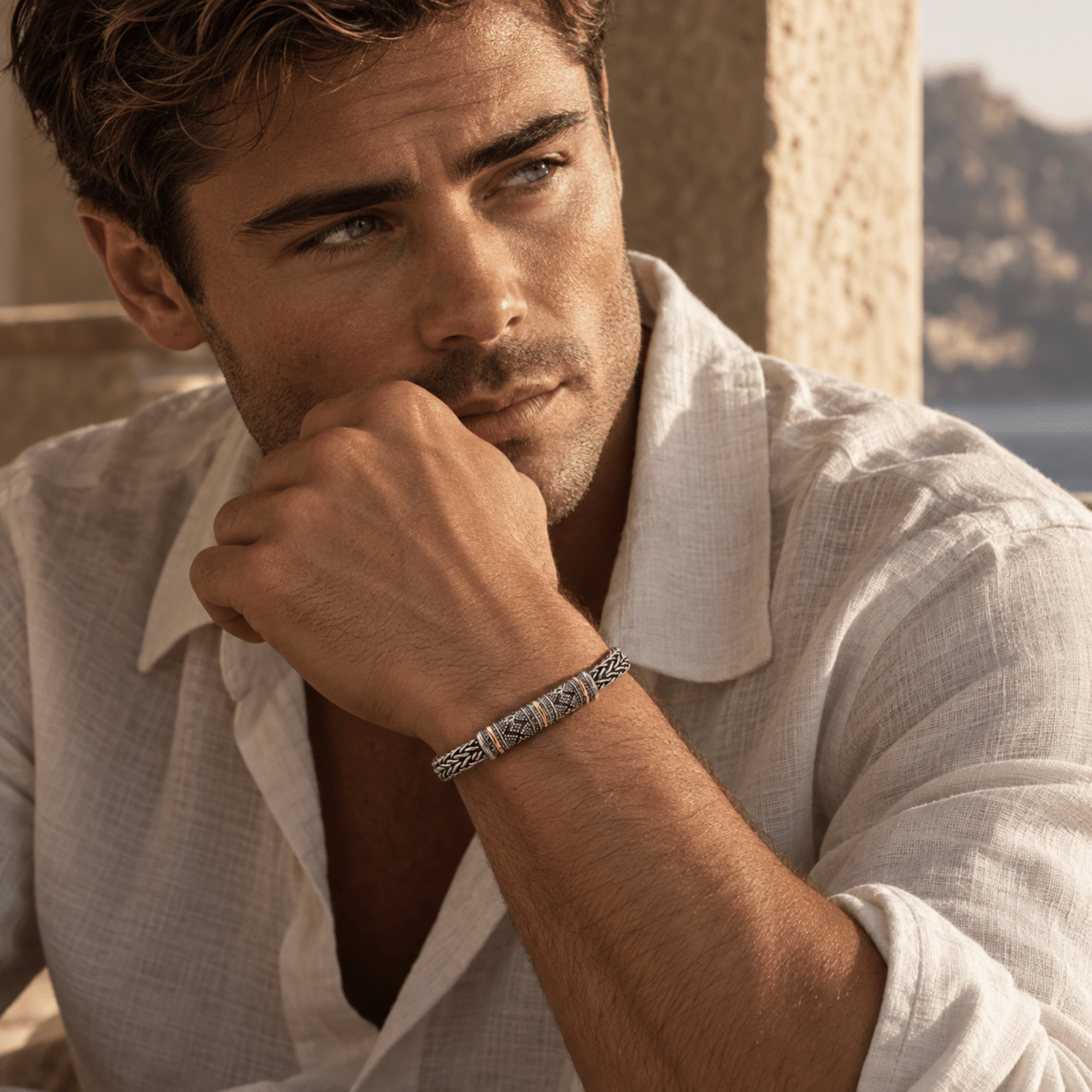 A man wearing a silver-toned metal bracelet with blue and gold geometric patterns, posing in a light-colored linen shirt against a warm stone background.