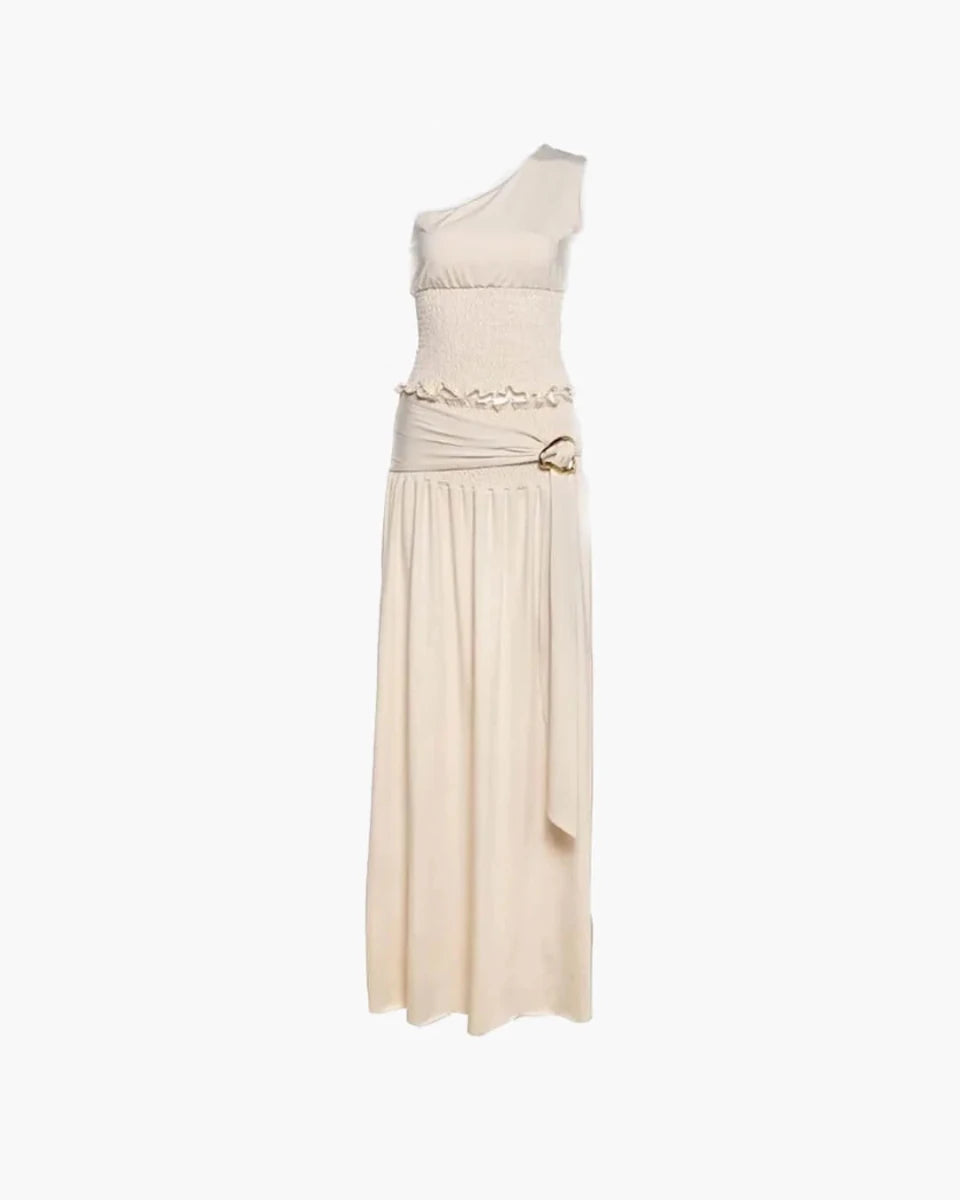 A one-shoulder maxi dress in a light cream or off-white color, featuring a textured bodice, a ruffled waistband with a gold-tone ring detail, and a flowing skirt, shown against a plain white background.