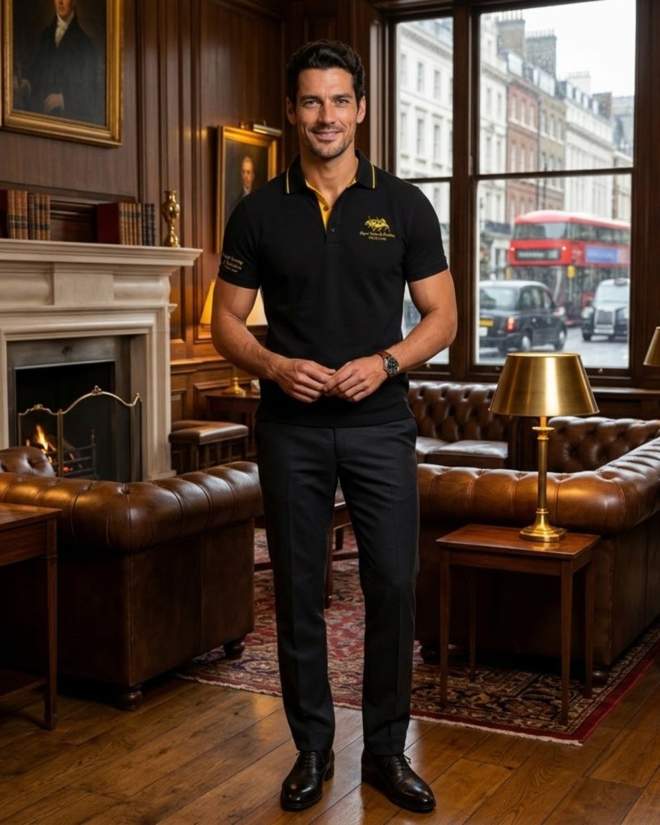 A man stands in a classic London pub wearing a black polo shirt with a yellow collar and embroidered logo, paired with black trousers. The background features a warm wood-paneled wall, a white fireplace, brown leather tufted sofas, and a window showing a red double-decker bus outside.