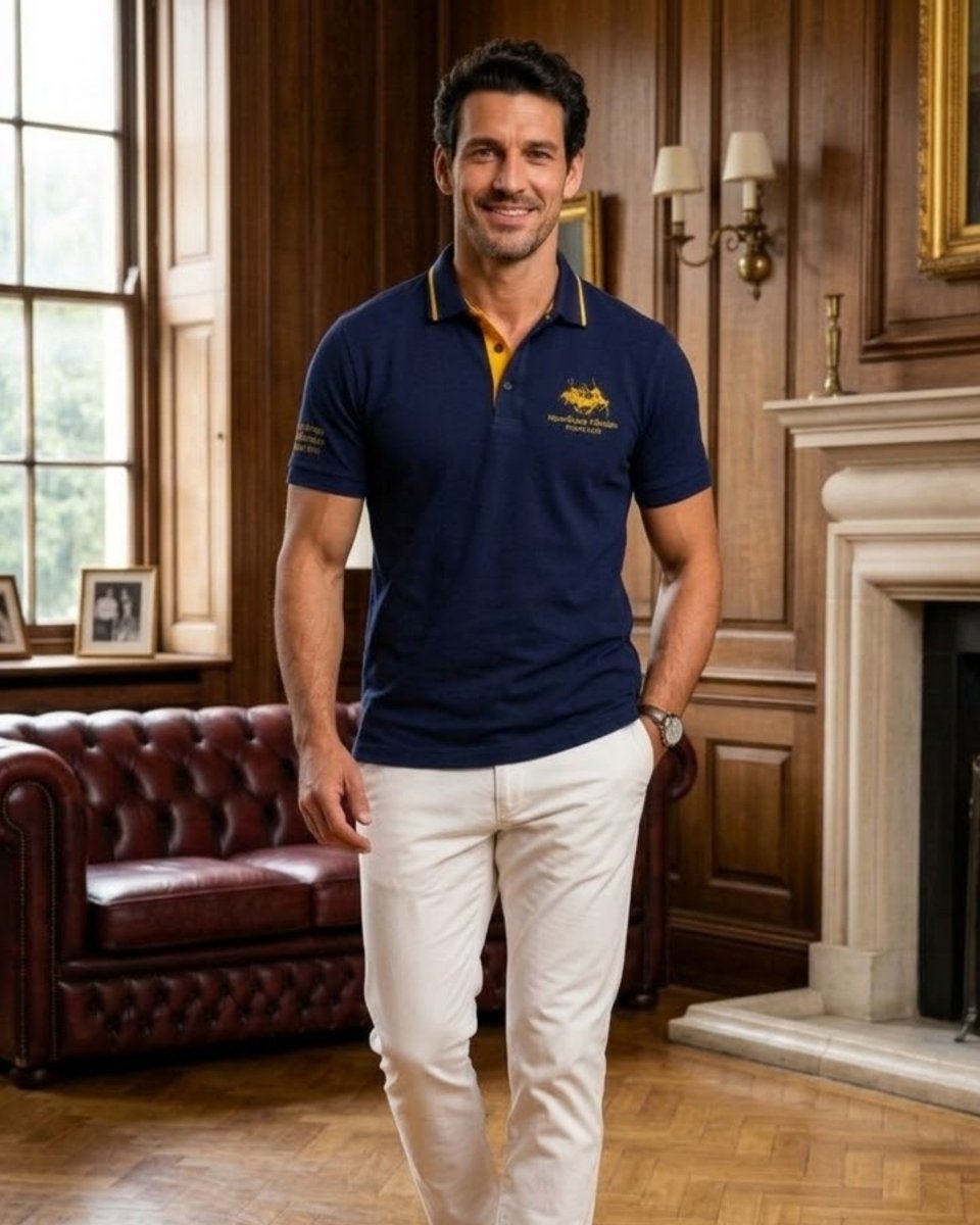 A man wearing a navy blue polo shirt with yellow accents and white trousers, standing in a classic wood-paneled room with a leather sofa and fireplace.