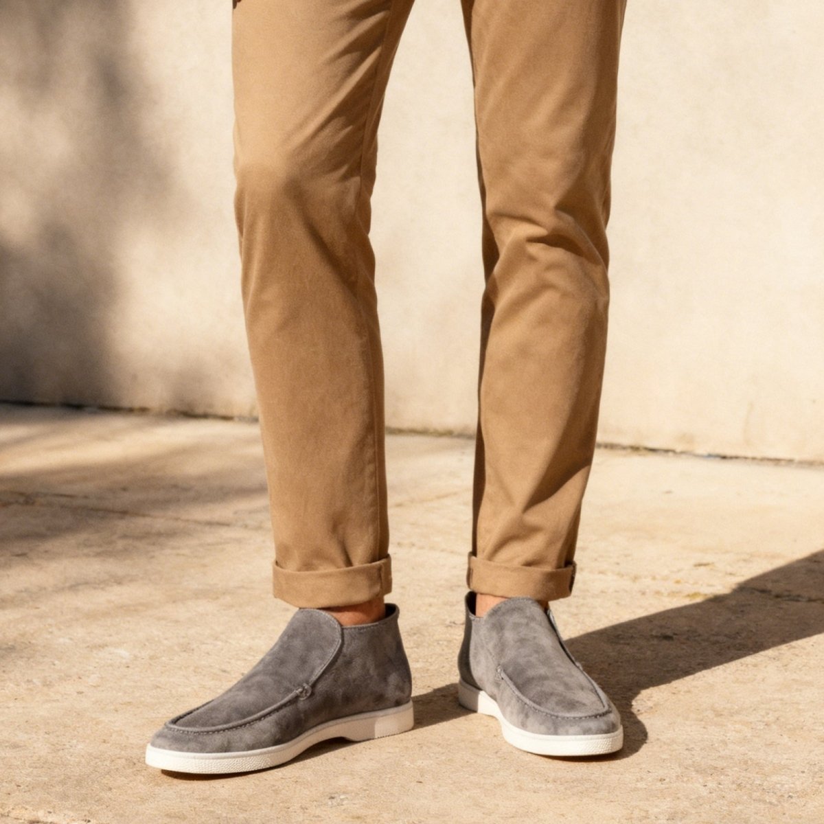 A man wearing Kouvr Fashion's grey suede loafer ralph boots with cuffed brown chino trousers, standing on a sunlit pavement.