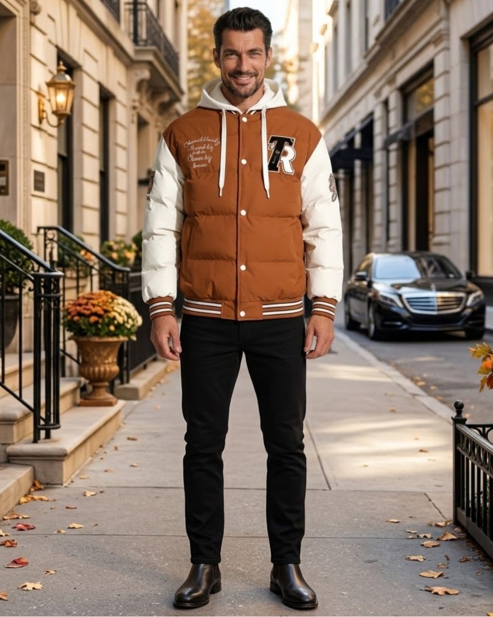 A man stands on a city sidewalk wearing a brown and cream varsity-style puffer jacket with a white hood, black pants, and dark brown boots. The background shows a real urban street scene with buildings, a parked car, and fallen leaves.