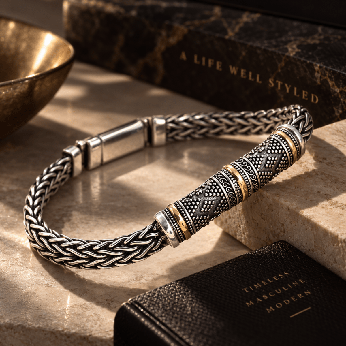 A silver-toned braided bracelet with a cylindrical textured centerpiece, accented by gold and dark oxidized details, displayed on a stone surface with a marble book and a black leather book in the background.