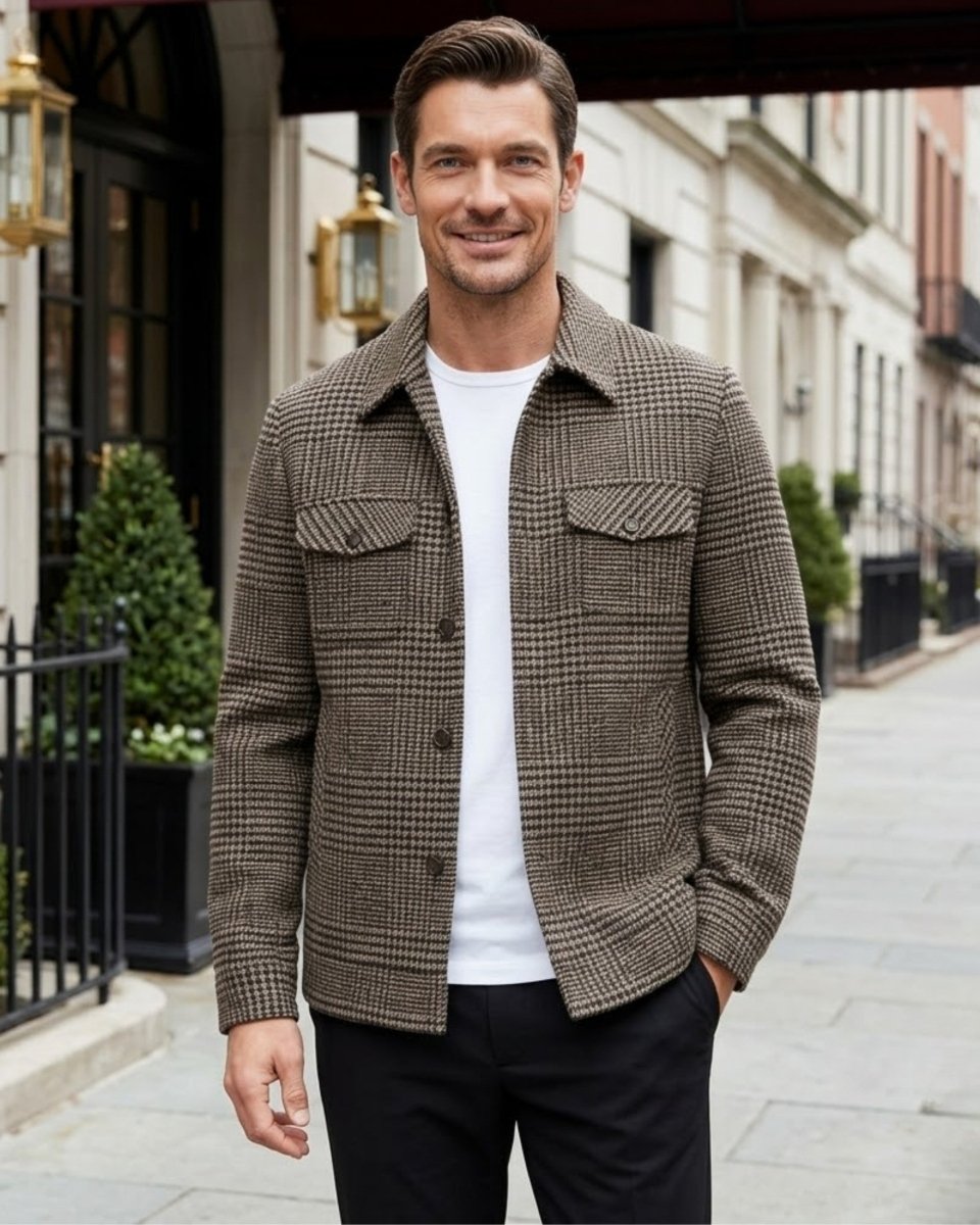 A man wearing a brown and beige houndstooth patterned button-up shirt jacket over a white t-shirt and black trousers, standing on a city street.