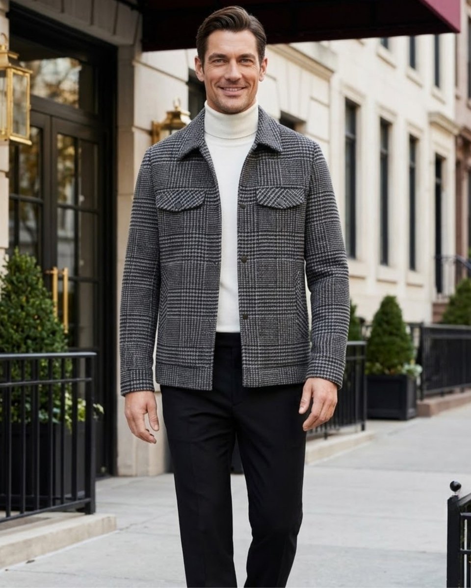 A man wearing a gray and black plaid button-down jacket over a white turtleneck sweater and black trousers, walking on a city sidewalk.