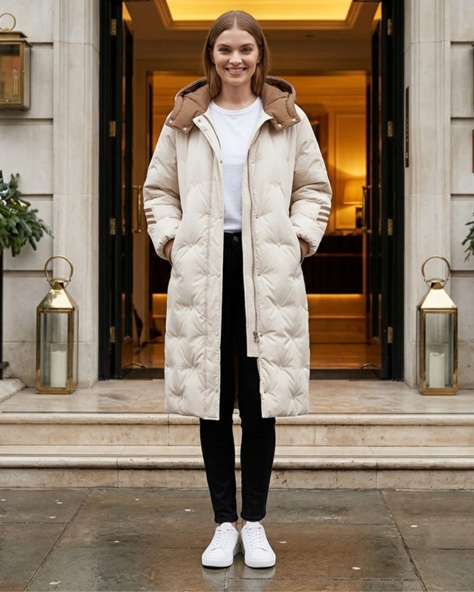 A woman stands in front of a building entrance wearing a long quilted puffer coat in a light cream color with a brown collar, over a white top and black pants.