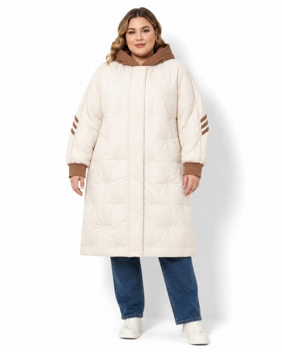 A woman modeling a long quilted puffer coat in cream with brown trim and hood, paired with blue jeans and white sneakers, against a plain white background.