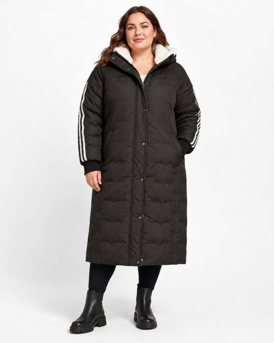 A woman modeling a long black puffer coat with a white sherpa-lined hood and white stripes on the sleeves, standing against a light gray studio background.