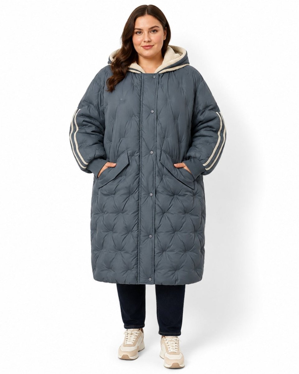 A woman modeling a long, dark slate gray quilted puffer coat with a cream-colored hood lining and cream stripes on the sleeves, paired with dark pants and beige sneakers, against a plain white background.