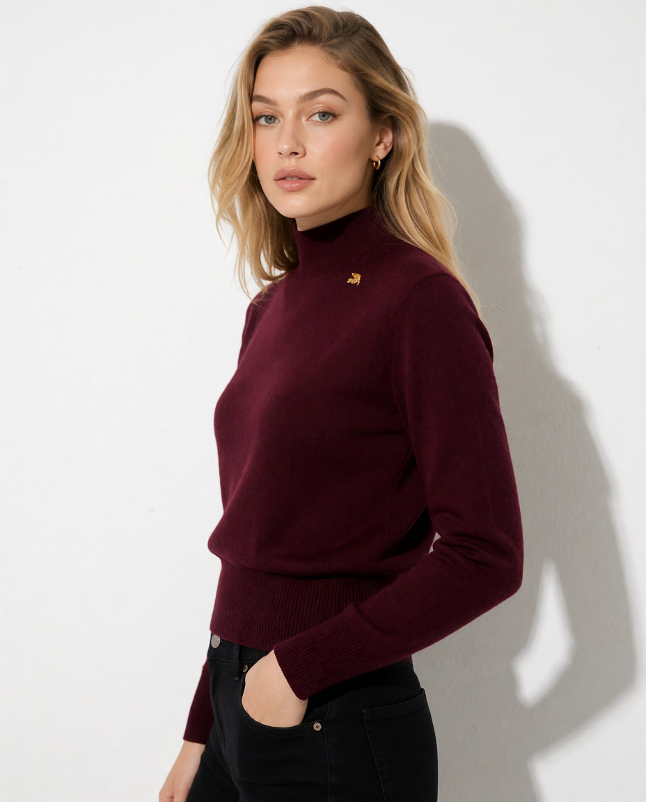 A woman wearing a long-sleeve ribbed turtleneck sweater in deep burgundy with a small gold embroidered logo on the chest, paired with black pants, standing against a white wall with a soft shadow.