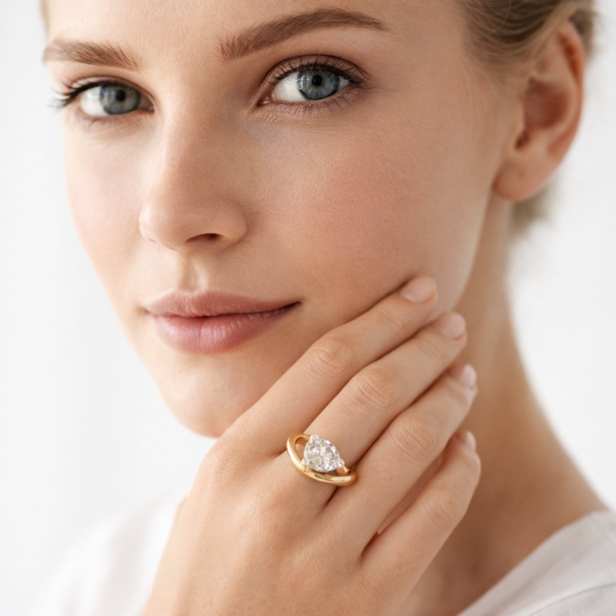 A woman with blue eyes wearing the Kouvr Zuri modern pear shaped diamond solitaire ring in yellow gold, touching her cheek.