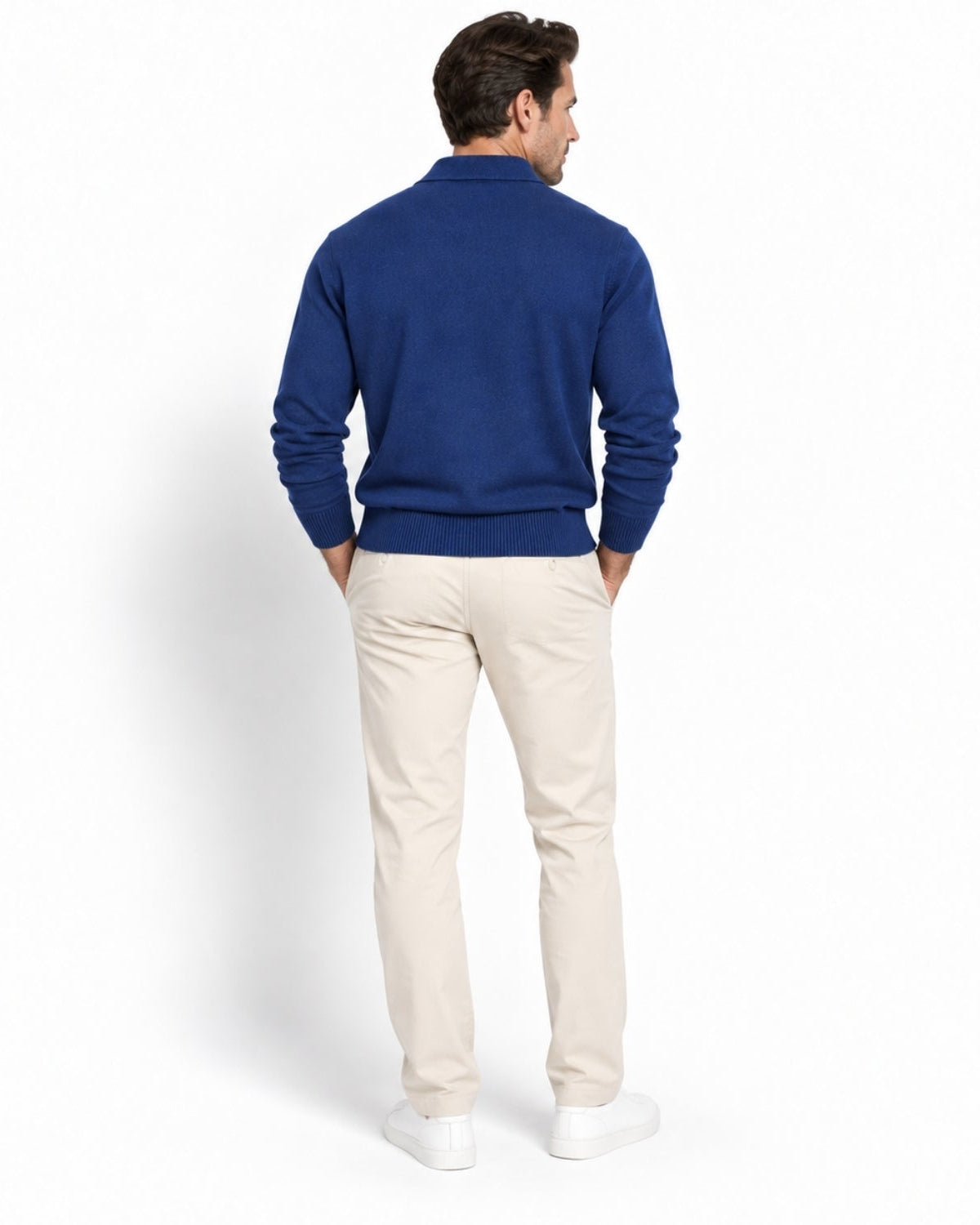 A man shown from the back wearing a royal blue long-sleeve knit sweater, beige trousers, and white sneakers, standing against a clean white studio background.