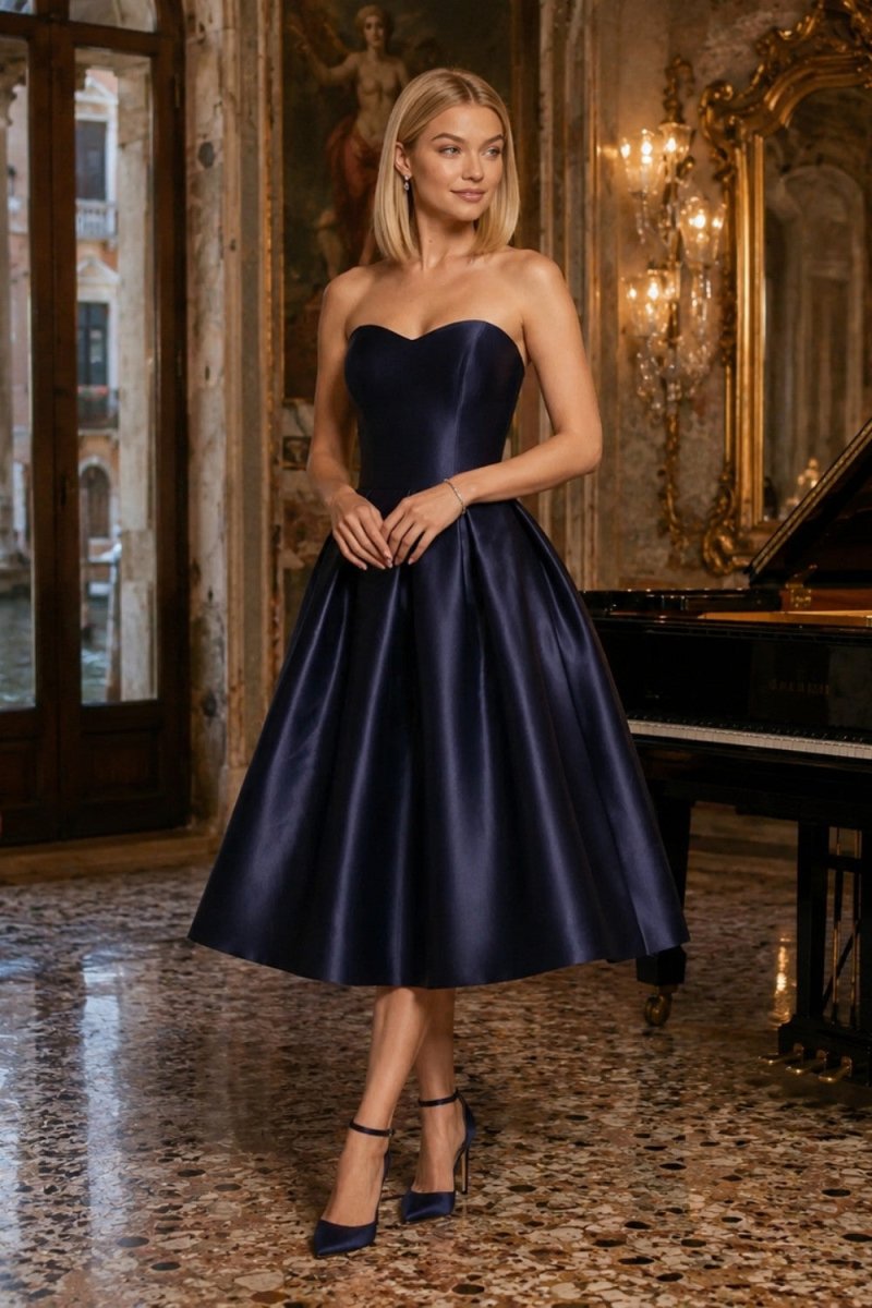 A woman models a strapless sweetheart A-line midi dress in dark navy blue satin, standing in an elegant, classic Italian-style room with a grand piano and chandeliers.