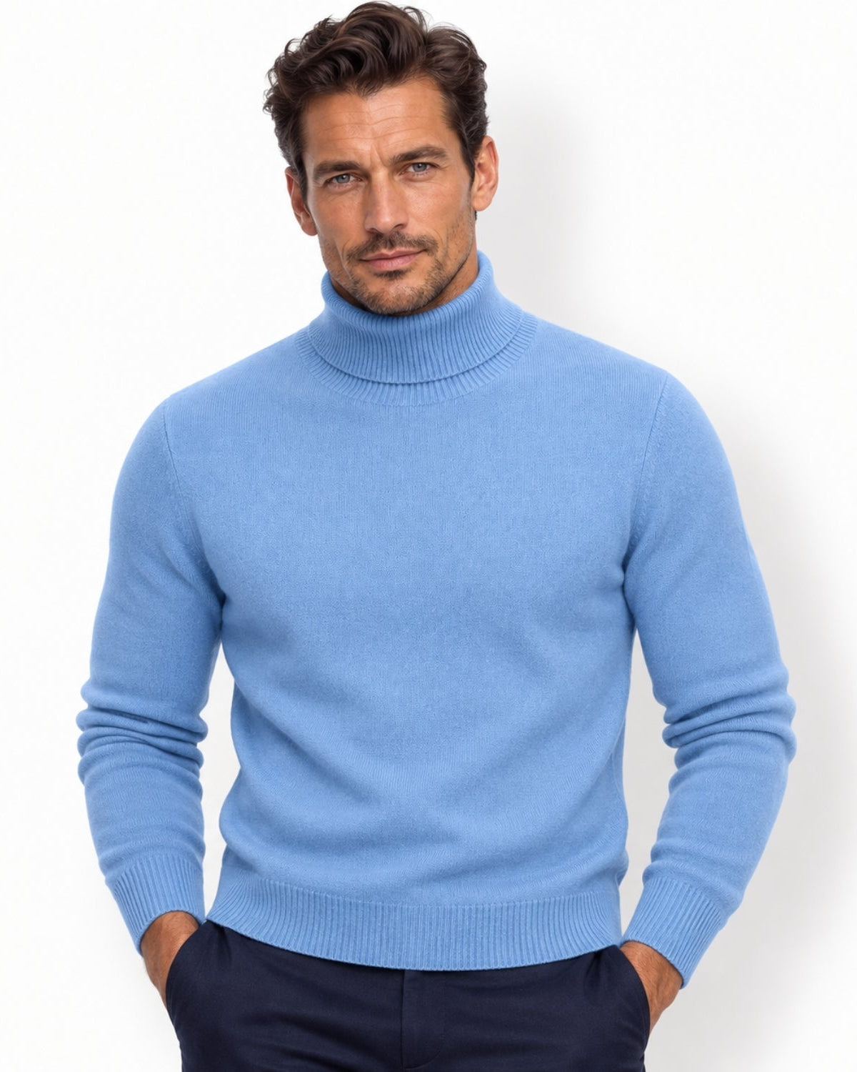 A man wearing a light navy knit turtleneck sweater and dark navy trousers, standing against a plain white studio background.