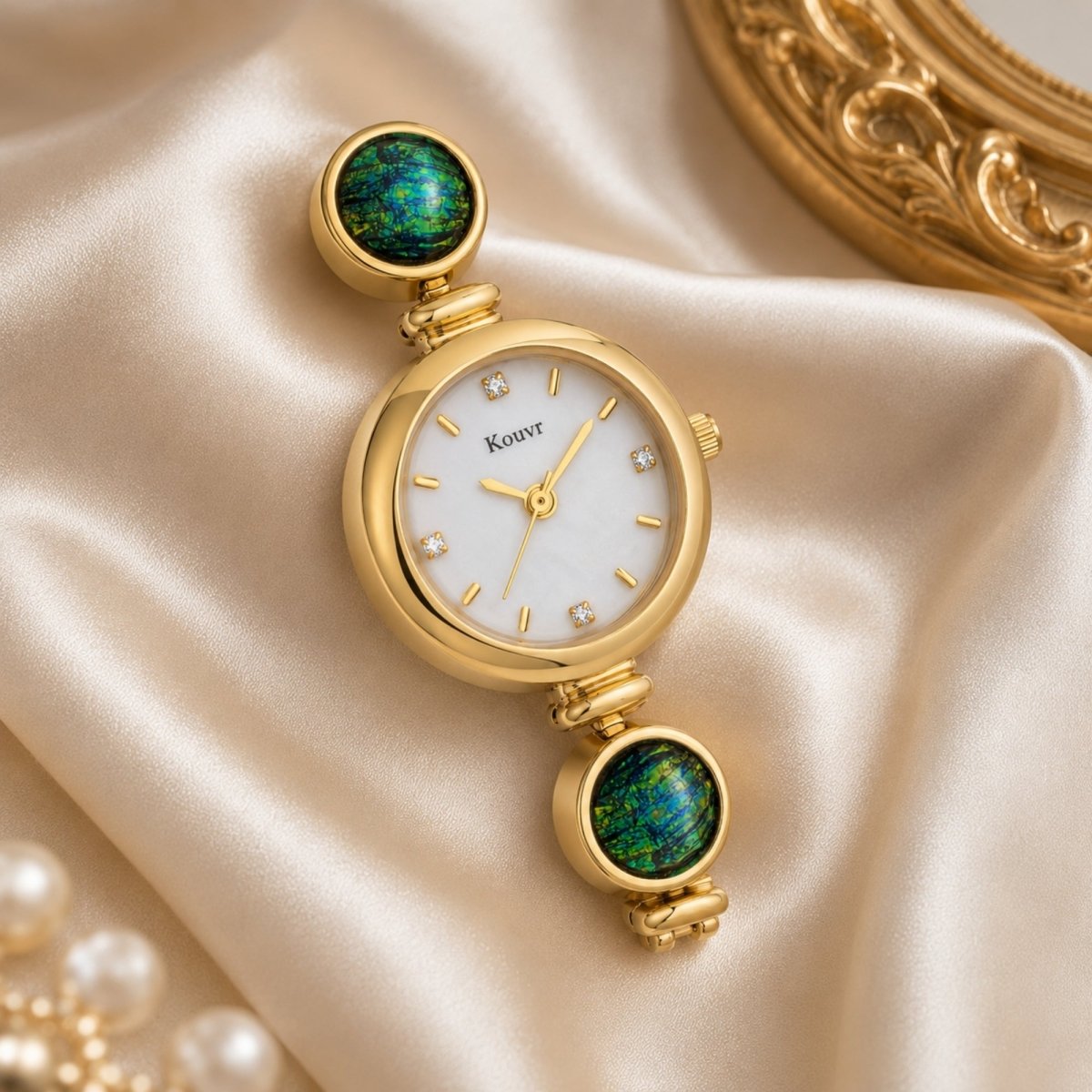 Elegant Kouvr gold women's watch with bright green stones resting on smooth champagne-colored silk fabric next to an ornate gold mirror frame.