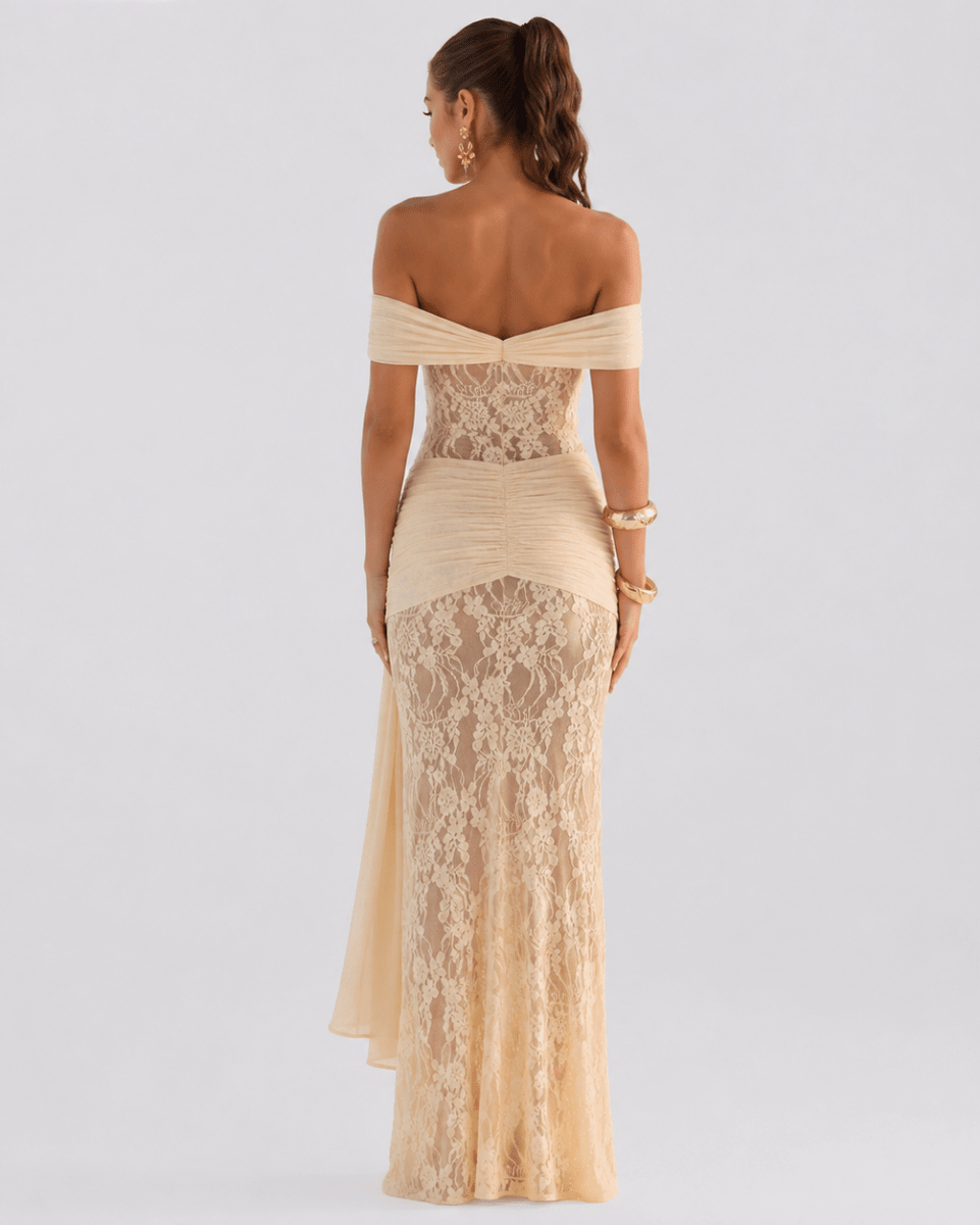 A woman viewed from the back wearing an off-the-shoulder formal gown in a warm champagne beige color with a sheer floral lace overlay and ruched detailing, set against a plain light gray studio background.