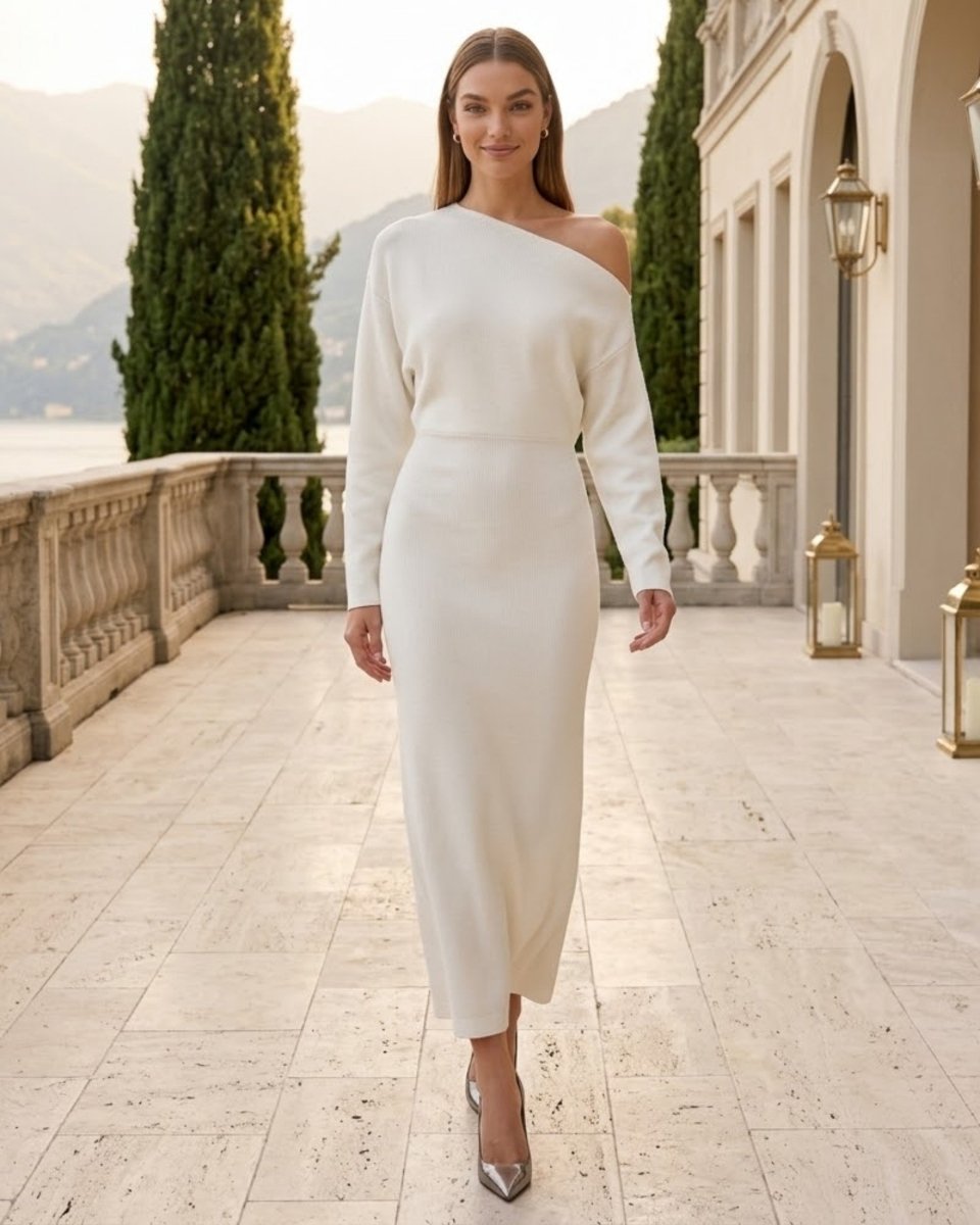 A woman models a long-sleeve, off-the-shoulder midi dress in a warm off-white or cream color, set against a luxurious outdoor terrace with stone balustrades, cypress trees, and a distant lake or sea.