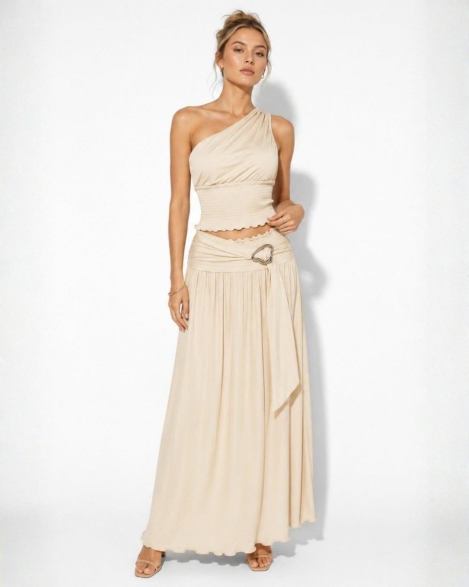 A woman modeling a two-piece beige outfit consisting of a one-shoulder crop top and a matching pleated maxi skirt with a decorative belt buckle, set against a plain white studio background.