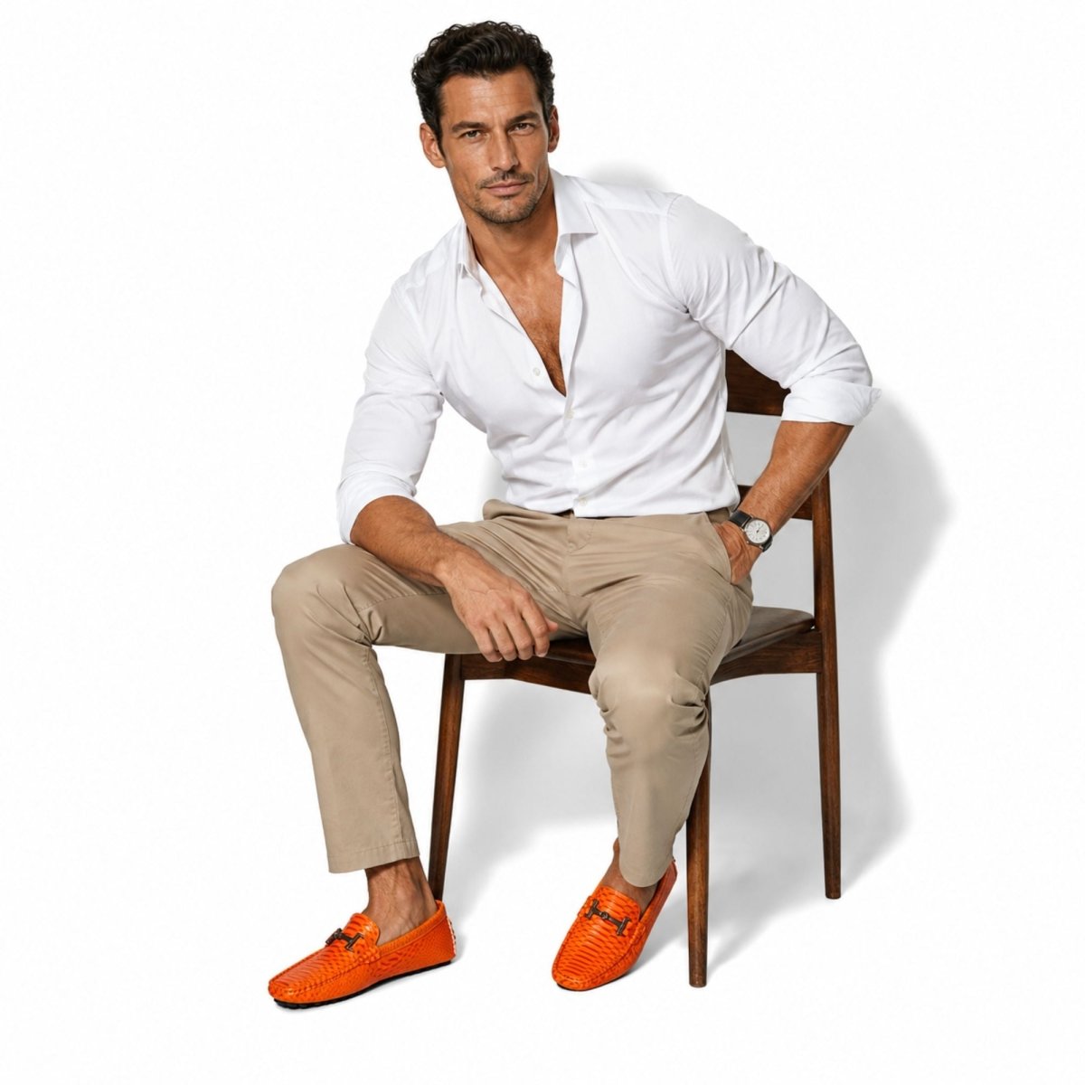 A man sits on a wooden chair wearing a white button-down shirt, beige trousers, and bright orange woven loafers against a clean white background.