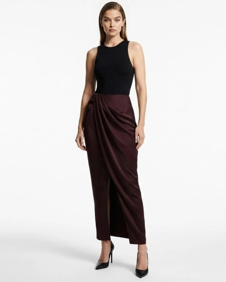 Model wearing Ourania high-waist draped sarong pants in deep aubergine satin, styled with a black tank top and heels.
