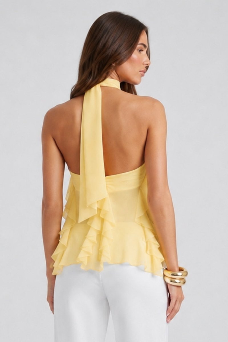 A woman shown from the back wearing a pale yellow ruffled halter-style top with a draped neck scarf detail and white pants, against a light gray studio background.