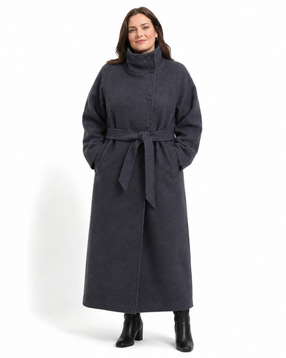 A woman modeling a dark charcoal gray belted coat with a mock neck and front buttons, paired with black ankle boots, against a plain white studio background.