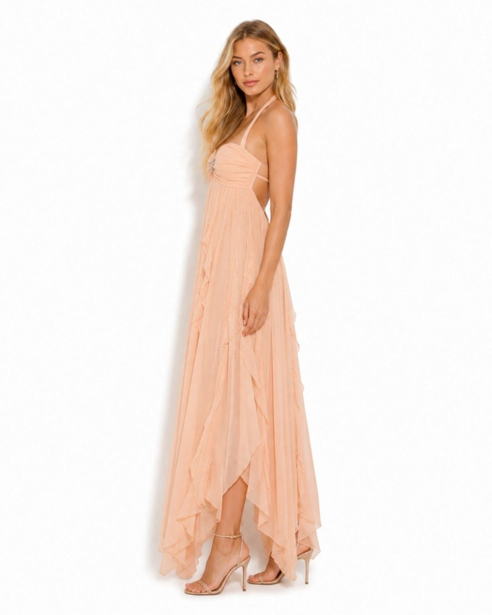 A woman modeling a peach-colored, floor-length halter dress with a ruffled, tiered skirt and a high-low hemline, paired with nude high-heeled sandals against a plain white background.