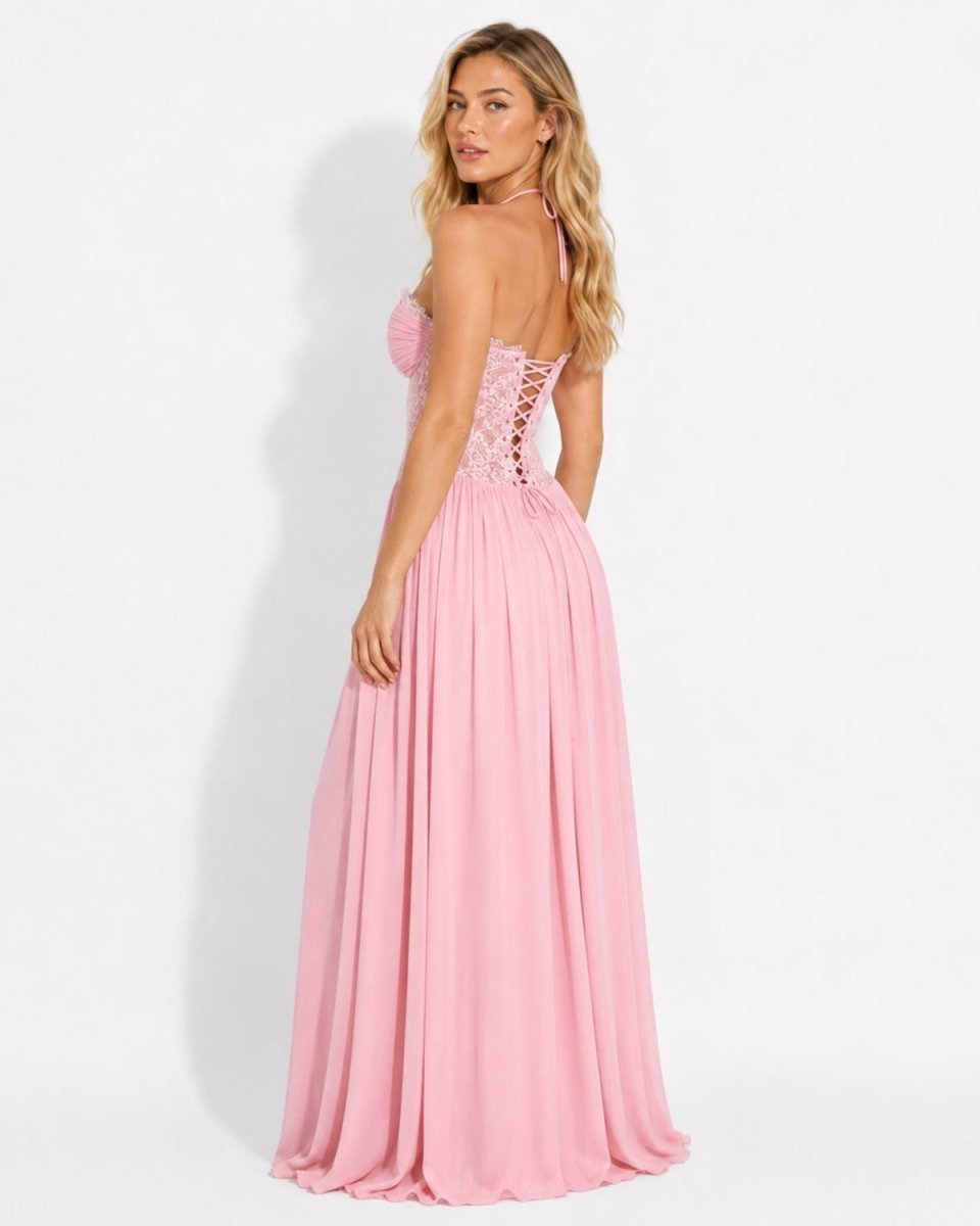 A woman modeling a long pink strapless gown with a lace-up back and flowing skirt, photographed against a plain light gray studio background.