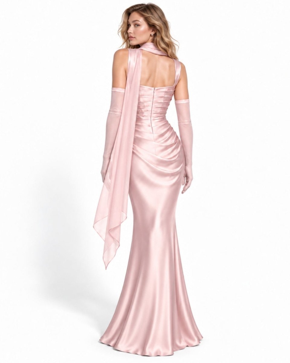 A woman modeling a light pink satin evening gown from the back, featuring a halter-style back, structured waist, long sheer gloves, and a matching draped scarf, set against a plain white studio background.