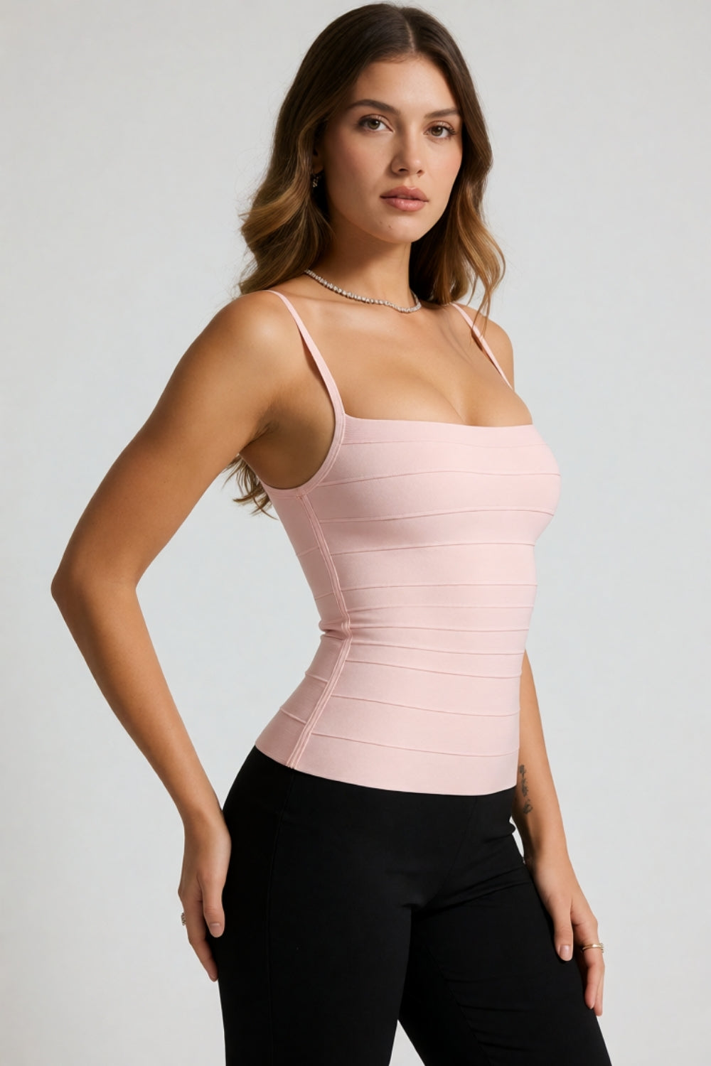 A woman modeling a light pink ribbed camisole top with thin straps and a square neckline, paired with black pants, against a light gray studio background.