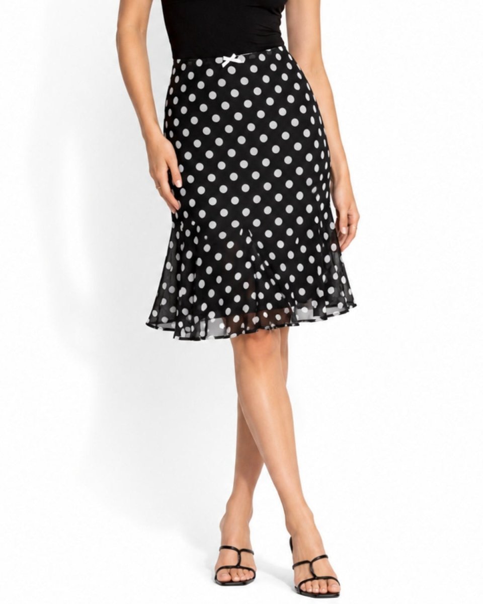 A woman models a black midi skirt with a white polka dot pattern, paired with a black top and black strappy sandals, against a plain white studio background.