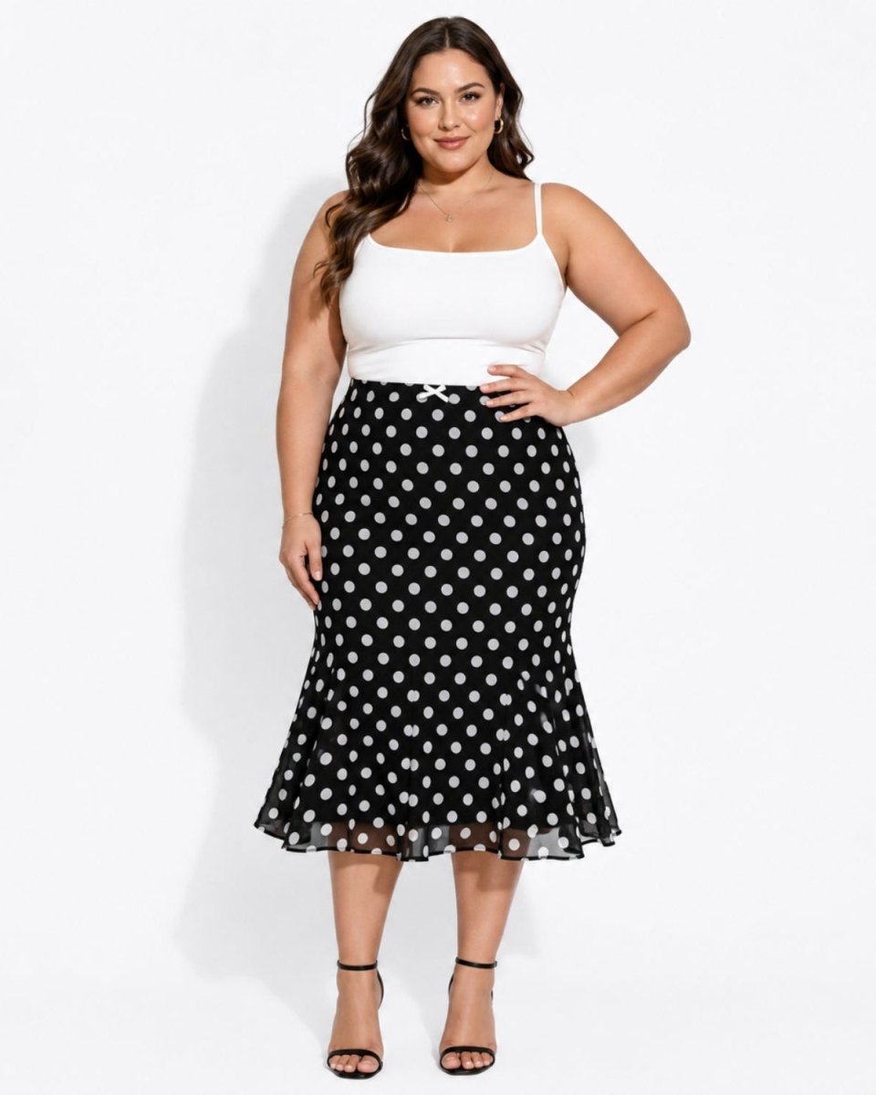 A woman modeling a black midi skirt with a white polka dot pattern, paired with a white spaghetti-strap top and black high-heeled sandals, against a plain white studio background.