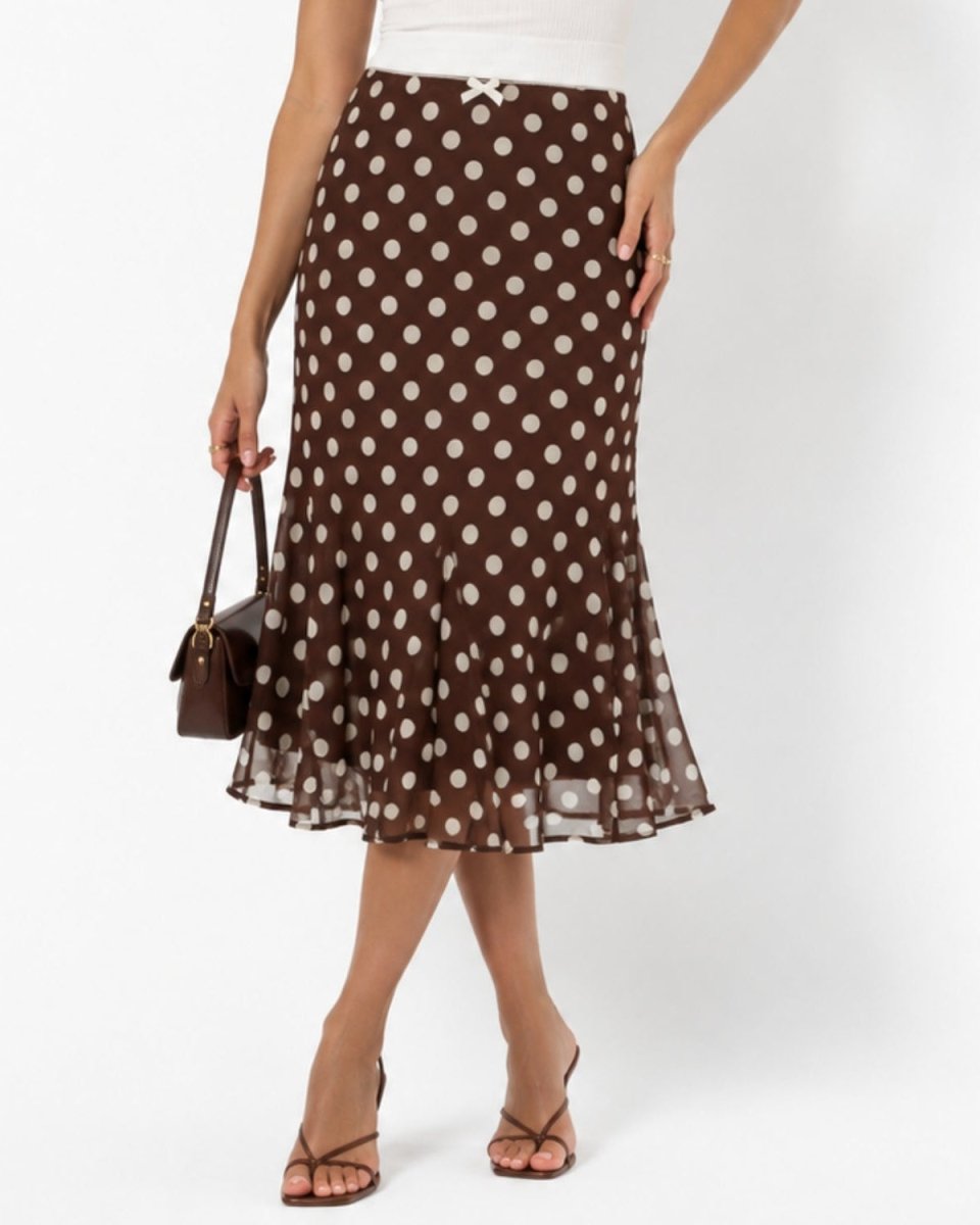 A model wearing a brown midi skirt with white polka dots, styled with a white top, brown strappy heels, and a small brown handbag against a plain white studio background.